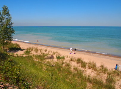 [View of the lake shore as seen from above. The hillside with plenty of green vegetation is in the foreground and several people are on the beach in the mid-ground. The lake with the Chicago haze above it is in the background.]